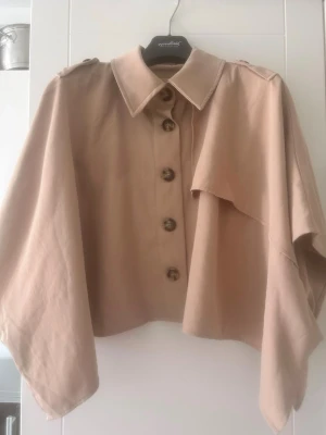 Kort beige trenchcoat med capeärm - Stilren kort trenchcoat i beige med stora knappar framtill och cape-liknande ärmar. Klassisk krage och axelklaffar ger en trendig look. Perfekt för dig som vill ha en unik twist på den klassiska trenchcoaten.