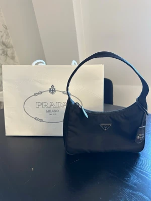Prada väska Re-edition nylon  - Svart handväska från Prada Re-edition 2000 nylon. Sparsamt använd. Finns några små dova fläckar på baksidan som jag ej testat rengöra.  Medföljer  -påse -äkthetsbevis -dustbag  Inköpt på Pradas hemsida och kostar idag 11 500kr.