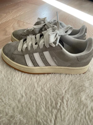 Grå Adidas Campus sneakers - Säljer grå Adidas Campus sneakers i mocka med vita ränder på sidan. Skorna har lågt skaft och breda vita skosnören.  
