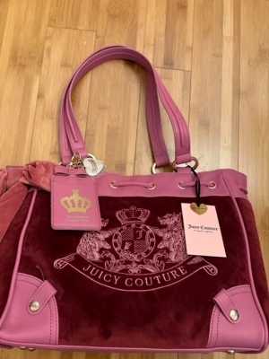 Juicy Couture daydreamer rosa och vinröd handväska - Juicy Couture handväska i rosa och vinröd sammet med detaljer i rosa konstläder. Väskan har dubbla handtag, guldfärgade metalldetaljer, emblem och logga framtill samt en söt tag med krona. Perfekt för dig som gillar statement-accessoarer!