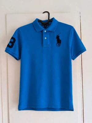 Ralph Lauren blå piké – Big Pony – storlek M (10-12) - Snygg blå Ralph Lauren-piké i storlek M (10-12) 💙 Klassisk modell med stor broderad Big Pony-logga på bröstet och nummer 3 på ärmen. Kortärmad med krage och knäppning framtill. Ser ut att vara i fint begagnat skick på bilderna. Perfekt till sommaren eller för en klassisk preppy/look. Helt okej skick men har några defekter! 📏 Storlek: M (10-12) 🎨 Färg: Klarblå med mörkblå detaljer 🐎 Ralph Lauren Big Pony