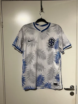 Vit Brasilien fotbollströja Nike - Snygg vit fotbollströja från Nike med Brasilien-emblem på bröstet. Tröjan har blå och grå palmbladsmönster, blå detaljer vid krage och ärmslut samt klassisk v-ringning. Perfekt för dig som älskar fotboll och vill sticka ut på planen.