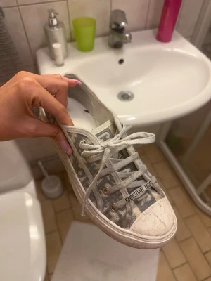 Gråa Dior sneakers med monogram - Säljer ett par gråa sneakers från Christian Dior med klassiskt monogrammönster och vita snören. Skorna har vit sula och detaljer med logotyp på sidan och framtill. Perfekta för dig som vill ha en lyxig och trendig look.