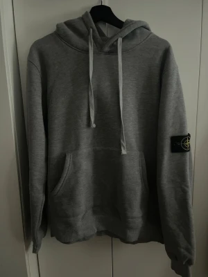 Grå hoodie med Stone Island patch - Säljer en grå hoodie från Stone Island med klassisk patch på vänster ärm. Hoodien har huva med dragsko och stor magficka framtill. Tillverkad i mjukt sweatshirtmaterial som är skönt att bära. Perfekt för en avslappnad och stilren look.