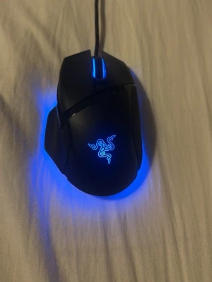 Svart Razor gamingmus med RGB-belysning - Säljer en svart gamingmus från Razer med RGB-belysning som kan ändra färg mellan blått och vitt. Musen har en ergonomisk form, flera sidoknappar och en sladd för anslutning. Perfekt för gaming och har den klassiska Razer-loggan som lyser upp.