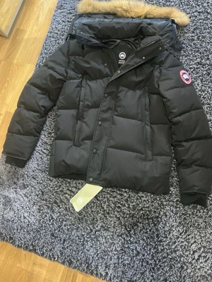 Svart dunjacka från Canada Goose - Säljer en svart dunjacka från Canada Goose med avtagbar huva och beige fuskpäls. Jackan har en klassisk patch på ärmen, dragkedja och knappar framtill samt ribbade muddar vid ärmsluten. Perfekt för kalla vinterdagar.