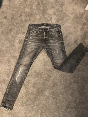 Grå jeans från Replay med slitningar - Säljer ett par grå jeans från Replay med slitningar och tvättade detaljer. Modellen är smal med klassisk femficksdesign och dragkedjegylf. Jeansen har en cool, använd look och passar perfekt till en avslappnad stil. [pris kan diskuteras vid snabb affär]