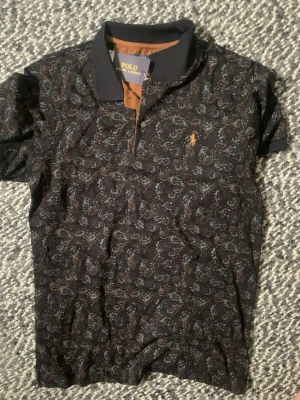 Mönstrad piké från Polo Ralph Lauren - Säljer en svart pikétröja från Polo Ralph Lauren med brunt och blått blommönster. Klassisk krage, knappslå och den ikoniska broderade loggan på bröstet. Perfekt för dig som vill ha en unik och stilren skjorta.