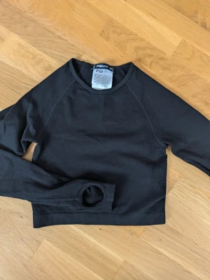 Svart långärmad träningströja - Svart långärmad träningströja med rund hals och tumhål i ärmarna. Tröjan har diskreta meshdetaljer längs sidorna och är tillverkad i ett stretchigt funktionsmaterial som andas. Perfekt för gymmet eller löprundan.