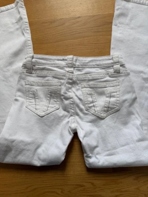 Lågmidjade jeans - Säljer ett par vita jeans med utsvängda ben och snygga broderade detaljer på bakfickorna. Klassisk femficksmodell med bälteshällor och knappgylf. Perfekta för dig som vill sticka ut med en retroinspirerad look.