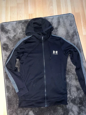 Svart hoodie från Under Armour - Svart zip-hoodie från Under Armour med gråa detaljer längs ärmarna och vit logga på bröstet. Tillverkad i ett mjukt och stretchigt funktionsmaterial, perfekt för träning eller chill. Huva med dragsko och hel dragkedja framtill.lite skadad vid framsidan  , men svårt att märka 