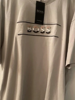 Beige t-shirt från BOSS med logga - Snygg beige t-shirt från BOSS med stor logga framtill i relief och två tunna linjer ovanför och under texten. Klassisk passform och korta ärmar, perfekt till jeans eller shorts för en clean look.