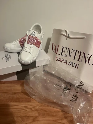 Valentino Garavani vita sneakers 37 - Snygga vita sneakers från Valentino Garavani med rosa och vita detaljer i mönstrad textil över vristen. Skorna har klassisk låg profil, vita snören och en robust gummisula med mönster. Perfekta för dig som vill sticka ut med exklusiv design. 37