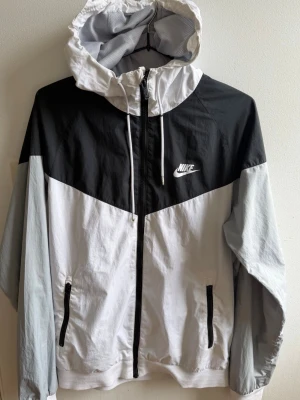 Nike svartvit vindjacka med huva - Nike vindjacka i svart, vitt och ljusgrått med dragkedja framtill, två fickor med dragkedja och justerbar huva. Jackan har en sportig look med färgblock och Nike-logga på bröstet. Tillverkad i lätt och vindtätt material, perfekt för blåsiga dagar.