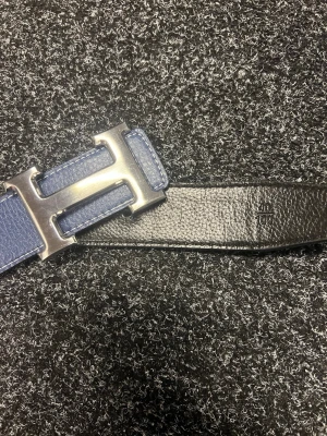 Blått och svart skärp med H-spänne - Snyggt skärp i blått och svart skinn med ett stort, silvrigt H-format spänne. Skärpet har en stilren och modern look, perfekt för att lyfta vilken outfit som helst. Materialet är präglat och ger en exklusiv känsla.