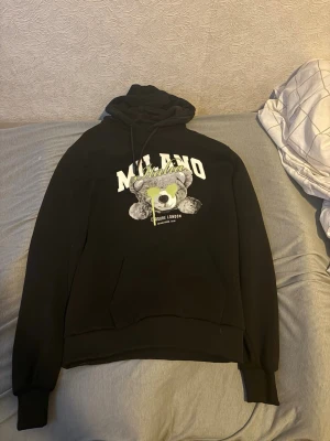 Closure london hoodie - Säljer en svart hoodie med huva och dragsnören. På bröstet står det 'MILANO' i vitt och ett stort tryck av en björn med texten 'SikSilk London'. Hoodien har magficka och ribbade muddar vid ärmar och nederkant. Perfekt för en chill och trendig look.