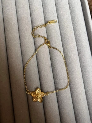 Guldfärgat armband med blomma - Snyggt armband i guldfärg med en detaljrik blomma i mitten. Kedjan är tunn och justerbar med en liten bricka märkt '18KGP', vilket betyder att det är guldpläterat. Perfekt för dig som gillar stilrena och feminina accessoarer.