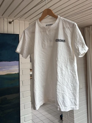 Jacquemus t-shirt - Säljer en vit t-shirt från Jacquemus med svart logotyptryck på bröstet och stor text på ryggen. Klassisk passform med rund hals och korta ärmar. Tillverkad i mjuk bomull som känns skön mot huden. Perfekt för dig som gillar stilrena och trendiga plagg.