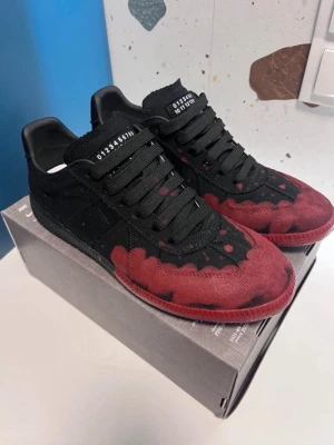Svarta och röda sneakers Maison Margiela - Unika sneakers från Maison Margiela i svart och rött med coolt färgstänksmönster. Skorna har svarta skosnören, låg profil och klassisk siluett. Materialet ser ut att vara canvas med detaljer i mocka och läder. Perfekta för dig som vill sticka ut.