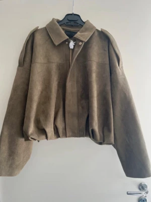 Populär zara mocka bomberjacka - Säljer en brun bomberjacka i mocka-look med krage och dragkedja framtill. Jackan har en loose passform och axelvaddar som ger en cool siluett. Perfekt för dig som vill ha en stilren och trendig jacka till höst och vinter.