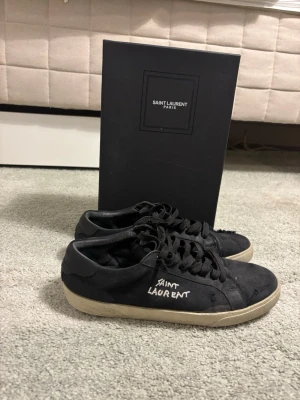 Svarta Saint Laurent sneakers - Svarta sneakers från Saint Laurent med vit logga broderad på sidan. Skorna har svarta skosnören och en vit sula som ger en snygg kontrast. Klassisk låg modell i canvas och mocka, perfekt för en trendig streetwear-look.