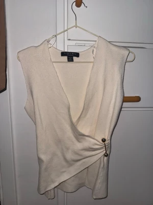Beige omlottopp  - Snygg beige omlottopp från Amisu med ärmlös design och v-ringad hals. Toppen har dekorativa guldfärgade knappar i sidan som ger en unik touch. Perfekt att styla med jeans eller kjol för en trendig look.