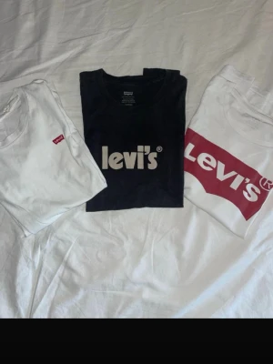 3-pack Levi's t-shirts med logga - Säljer ett 3-pack Levi's t-shirts i bomull. Två vita t-shirts, en med röd logga och en med liten röd patch, samt en svart t-shirt med stor vit Levi's-logga. Alla har rund hals och klassisk passform. Perfekta för en clean och stilren look.