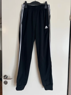 Svarta Adidas mjukisbyxor med ränder - Säljer ett par svarta mjukisbyxor från Adidas med klassiska vita ränder längs sidorna och Adidas-logga på låret. Byxorna har resår i midjan och vid bensluten, samt smal passform. Perfekta för chill eller träning. Har använts en del. Har tagit bilder på några ställen där det har blivit nopprigt. Finns kanske fler ställen det är så på. Går säkert att få bort med noppborttagare om man vill.   Midja: 32cm Längd: 98cm