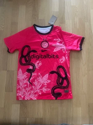 Inter Milan rosa matchtröja Nike - Säljer en rosa Inter Milan matchtröja från Nike med svart ormmotiv och vita bladmönster. Tröjan har korta ärmar, svart krage och muddar samt tryckt sponsorlogga på bröstet. Perfekt för dig som vill sticka ut på planen eller på läktaren.