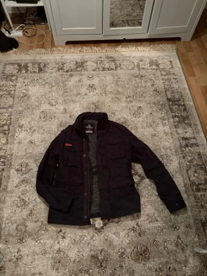 Svart vindjacka med fickor - Snygg svart vindjacka med fyra framfickor och hög krage. Jackan har dragkedja och knappar framtill samt diskreta röda detaljer. Perfekt för dig som vill ha en stilren och funktionell jacka till vardags.