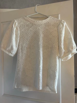 Vit spetsblus med puffärm - Säljer en söt vit blus med puffiga korta ärmar och rund hals. Blusen har ett detaljerat broderat mönster över hela plagget och ger en söt vibe. Perfekt att styla med jeans eller kjol för en trendig look.