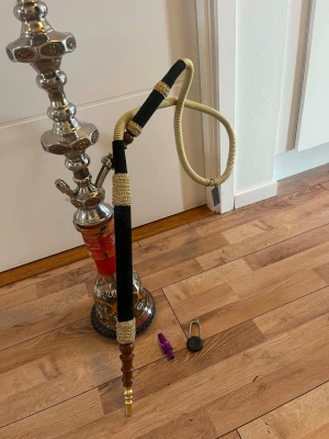 Stor vattenpipa med slang och tillbehör - Säljer en stor vattenpipa i silverfärgad metall och glas med dekorativa mönster. Medföljer en lång slang i svart och beige, samt tillbehör som koltång och munstycke. Perfekt för dig som vill ha en klassisk shisha till samlingen.