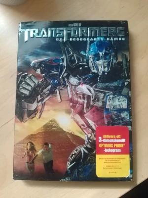 Transformers De besegrades hämnd DVD - DVD-film Transformers: De besegrades hämnd med omslag i färgstark design. Framsidan visar Optimus Prime och pyramider, samt en etikett om 3D-hologram. Filmen är regisserad av Michael Bay och texten är på svenska.