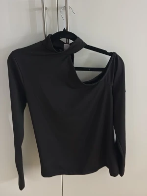 Svart långärmad one shoulder topp  - Svart långärmad one shoulder topp. Toppen har hög hals och är tillverkad i ett stretchigt material som ger en slim passform.