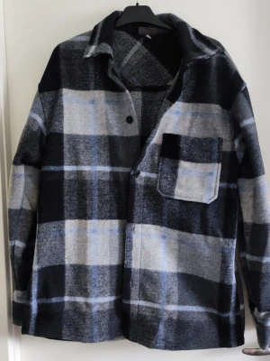 Rutig overshirt från H&M - Snygg overshirt från H&M i svart, grått och blått rutmönster. Skjortjackan har en bröstficka, klassisk krage och knäppning framtill. Tillverkad i mjukt, tjockt tyg som passar perfekt för lager på lager-stil.
