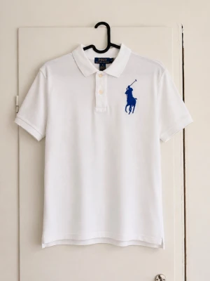 Ralph Lauren piké - Säljer en klassisk vit pikétröja från Polo Ralph Lauren med den ikoniska blå Big Pony-loggan på bröstet. Stilren och tidlös modell som passar perfekt till både vardag och sommar. ✔️ Original från Polo Ralph Lauren ✔️ Broderad Big Pony-logga ✔️ Fräsch vit färg ✔️ Bekväm passform och hög kvalitet.                                  Den är i bra skick men har en fläck!