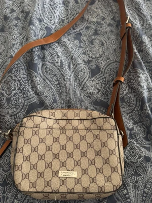 Beige axelväska från Don Donna - Snygg beige axelväska från Don Donna med brunt DG-mönster över hela väskan. Väskan har justerbar brun axelrem i skinnimitation och dragkedja upptill. Perfekt storlek för det viktigaste och en stilren detalj till din outfit.