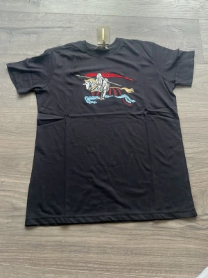 Svart Burberry t-shirt med tryck - Snygg svart t-shirt från Burberry med färgglatt tryck av en riddare på häst framtill. Tillverkad i mjuk bomull som känns skön mot huden. Klassisk passform och korta ärmar gör den enkel att matcha till olika outfits.