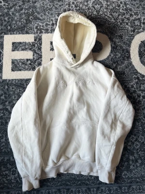 Vit hoodie Sweet Sktbs brodyr - Säljer en vit hoodie från Sweet Sktts med broderad logga på bröstet. Hoodien har en stor huva, magficka och ribbade muddar vid ärmar och nederkant. Tillverkad i mjukt bomullsmaterial för en skön och avslappnad look.