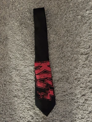 Svart slips med röd KISS-logga - Cool svart slips med stor röd KISS-logga i grafisk stil längst ner. Perfekt för dig som gillar rock och vill sticka ut med din accessoar. Slipsen har en klassisk form och är tillverkad i ett slätt tyg som ger en snygg finish. Helt ny. Ca 11 cm bred nertill.