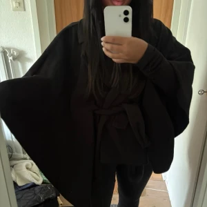 Svart cape-kappa med knytskärp - Säljer en svart cape-kappa med vid passform och knytskärp i midjan. Kappan har en elegant och flowy siluett, perfekt för lager på lager och en stilren look. Inga synliga knappar eller dragkedjor, och materialet ser ut att vara mjukt tyg.