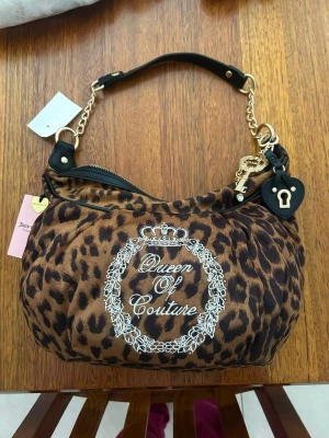 Leopardmönstrad handväska Queen of Couture - Snygg handväska med leopardmönster och broderad text 'Queen of Couture' i vitt. Väskan har guldfärgade kedjedetaljer, svart axelrem och dekorativa hängen i form av hjärta och nyckel. Materialet är textil med konstläderdetaljer. Perfekt för dig som gillar statement-accessoarer.