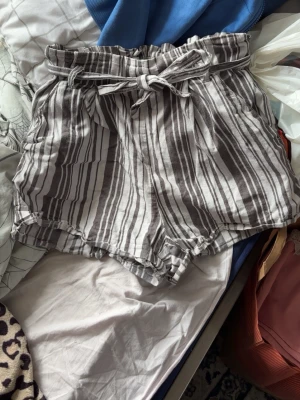 Randiga högmidjade shorts med knyt - Säljer ett par luftiga högmidjade shorts med grå-vita ränder och knytband i midjan. Shortsen har avslappnad passform och är perfekta för varma dagar. Tillverkade i ett mjukt och svalt material som känns skönt mot huden.