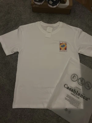 Vit t-shirt från Casablanca - Vit t-shirt från Casablanca med färgglatt tryck på bröstet där det står 'Casa Blanca'. Klassisk rund hals och korta ärmar. Perfekt för en clean och stilren look. Den är unik och passar perfekt till skolstarten, fraktar inom 0-1 dagar!