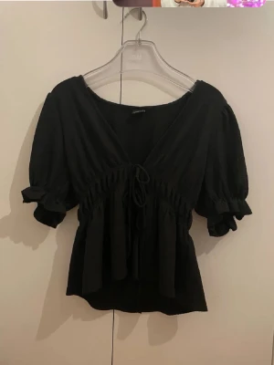 Svart blus med snörning Gina Tricot - Supersöt svart blus från Gina Tricot med v-ringning, puffärmar och snörning framtill. Blusen har volangdetaljer vid ärmslut och en smickrande passform som markerar midjan. Perfekt att styla med jeans eller kjol!