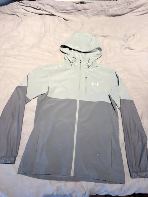 Grå vindjacka från Under Armour - Snygg tvåtonad grå vindjacka från Under Armour med huva och hel dragkedja. Jackan har en bröstficka med dragkedja, sidofickor och justerbar huva. Tillverkad i lätt och funktionellt material, perfekt för blåsiga dagar.