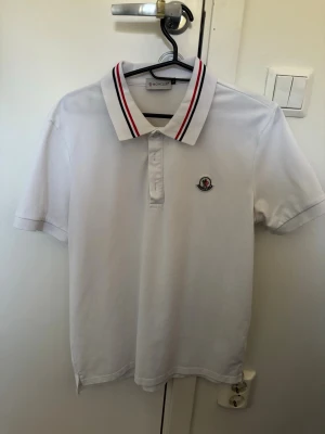  pikétröja Moncler - Klassisk vit pikétröja från Moncler med korta ärmar och krage med röda och blå ränder. Tröjan har Moncler-logga broderad på bröstet och knäppning framtill. Perfekt till en clean och sportig stil.