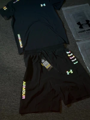 Svart träningsset från Under Armour - Snyggt svart träningsset från Under Armour med t-shirt och shorts. Båda plaggen har regnbågsfärgade logotyper och textdetaljer. Perfekt för gymmet eller löprundan. Materialet är lätt och andas, vilket gör det bekvämt under träning.