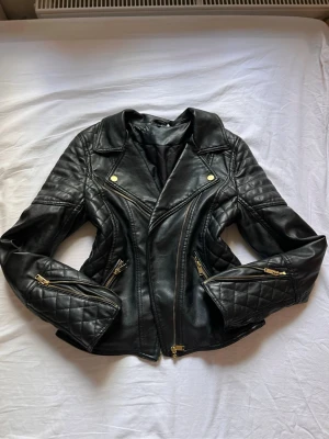 Svart biker skinnjacka med gulddetaljer - Snygg svart skinnjacka i bikerstil med guldfärgade dragkedjor och knappar. Jackan har quiltade detaljer på ärmarna och två framfickor med dragkedja. Snygg passform och klassisk krage ger en cool look.