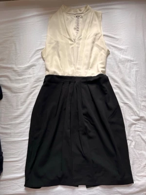 Svart och vit ärmlös klänning - Stilren ärmlös klänning med vit omlottdesign upptill och svart kjoldel. Klänningen har v-ringad hals och markerad midja, vilket ger en elegant siluett. Perfekt för dig som gillar klassiska färgkombinationer och enkel design.